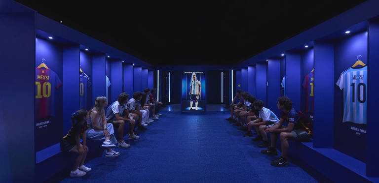 A imagem mostra uma sala de exibição com paredes azuis, onde várias camisas de futebol estão penduradas. À frente, um grupo de jovens sentados em bancos observa uma tela que exibe uma figura em uma camisa de futebol. As camisas penduradas incluem uma do Barcelona e outra da seleção argentina, ambas com o nome 'MESSI' e o número 10. A iluminação é suave, destacando as camisas e a tela.