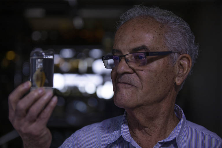 Um homem idoso com cabelo grisalho e óculos está segurando um pequeno objeto em uma mão， olhando para ele com uma expressão de curiosidade. O fundo é desfocado， com luzes suaves que sugerem um ambiente interno.