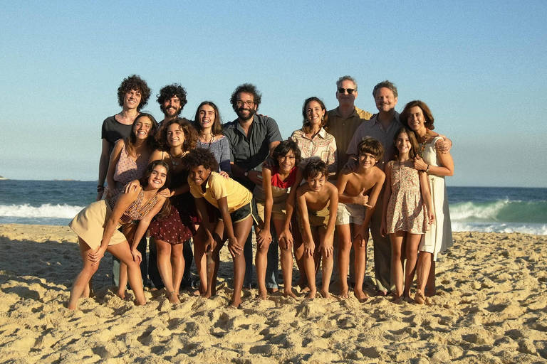 Uma foto em grupo tirada na praia, com um fundo de mar e céu claro. O grupo é composto por adultos e crianças, todos sorrindo. As pessoas estão vestidas de maneira casual, algumas com roupas de verão. O cenário é de areia e ondas ao fundo.