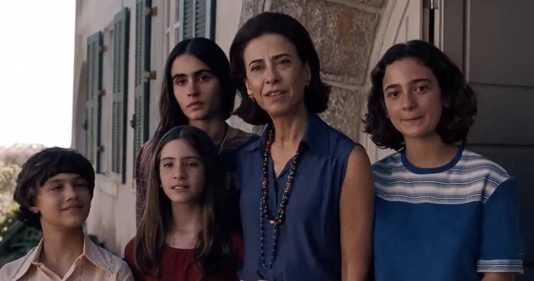 A imagem mostra um grupo de cinco pessoas em frente a uma casa. Há uma mulher adulta no centro, com cabelo escuro e preso, vestindo uma blusa azul e colar. À sua esquerda, uma menina com cabelo longo e liso, vestindo uma blusa vermelha. À direita, outra menina com cabelo curto e encaracolado, usando uma camiseta azul com listras. Atrás delas, um menino com cabelo castanho e uma menina com cabelo liso e escuro. O fundo apresenta uma casa com janelas verdes e uma parede de pedra.