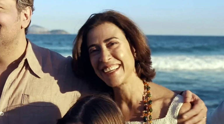 A imagem mostra uma mulher sorridente em primeiro plano， com cabelo castanho e liso， usando uma blusa clara e um colar colorido. Ela está ao lado de um homem e uma criança， todos posando juntos em uma praia， com o mar ao fundo e ondas visíveis. O céu está claro e ensolarado.