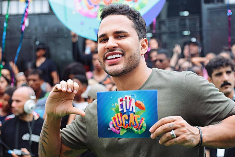 Um homem sorridente está segurando um cartaz colorido que diz 'Eita Lucas!' em um evento ao ar livre. Ele está vestido com uma camiseta verde e tem um anel em uma das mãos. Ao fundo, há uma multidão de pessoas, algumas segurando bandeiras e outros cartazes. O ambiente é festivo, com decorações coloridas e balões visíveis.