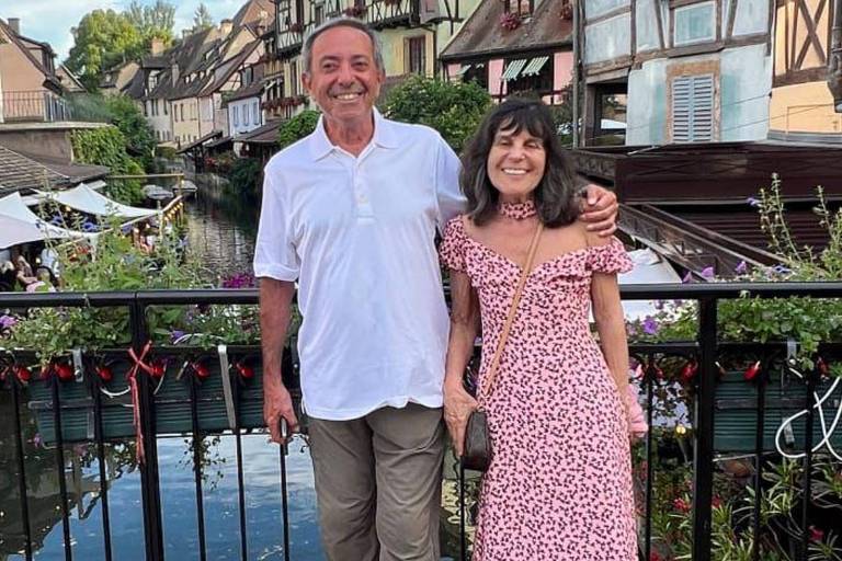 Um casal posando em uma vila pitoresca, com casas de estilo tradicional ao fundo. O homem está vestindo uma camisa branca e calças claras, enquanto a mulher usa um vestido rosa. Eles estão em frente a um pequeno canal, com mesas ao ar livre visíveis nas laterais. O céu está claro com algumas nuvens.