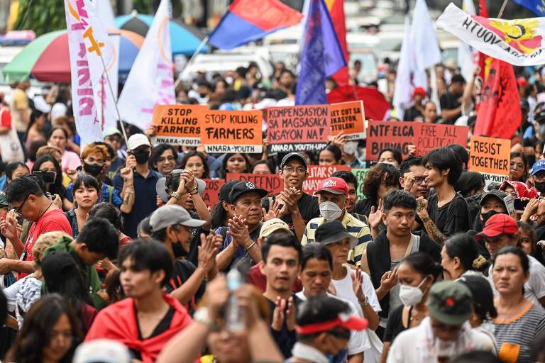 A imagem mostra uma grande multidão participando de um protesto. Muitas pessoas seguram cartazes com mensagens como 'STOP KILLING FARMERS' e 'COMPENSATION FOR MAGKUKULANGAN'. A cena é vibrante, com bandeiras coloridas ao fundo e um clima de mobilização. As pessoas estão vestidas de maneira variada, algumas usando chapéus e outras com roupas em tons escuros.