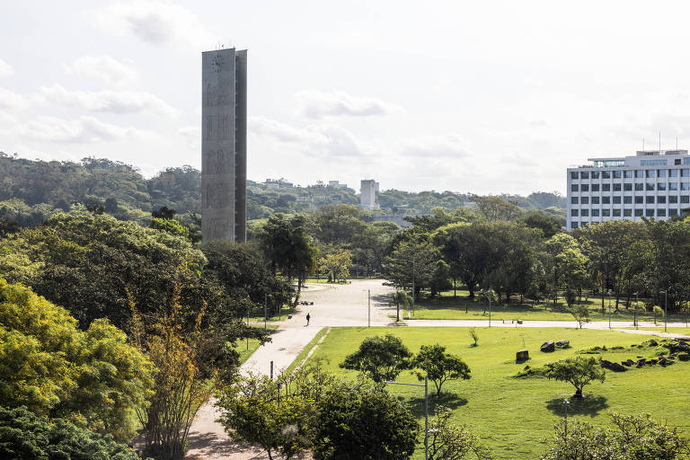 A imagem mostra uma vista de um parque com uma torre alta ao fundo. A torre possui um relógio e está cercada por árvores verdes. No primeiro plano， há um gramado amplo e algumas pessoas caminhando. O céu está parcialmente nublado， e há edifícios ao fundo， incluindo um prédio de escritórios.