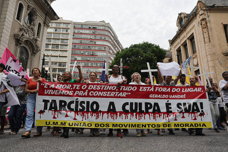 A imagem mostra um grupo de pessoas participando de uma manifestação. Elas seguram uma faixa grande com os dizeres 039;TARCÍSIO， A CULPA É SUA!039; e outras mensagens relacionadas à moradia. Algumas pessoas estão segurando cruzes brancas. O ambiente urbano ao fundo apresenta prédios e uma atmosfera de protesto.