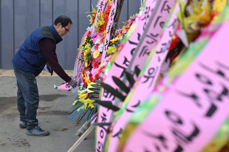 Um homem está se curvando para observar faixas coloridas com flores. As faixas são predominantemente rosas e possuem inscrições em caracteres coreanos. O ambiente parece ser ao ar livre, com um fundo cinza e um piso de terra.