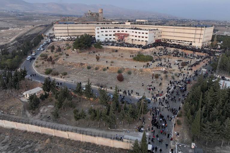 A imagem mostra uma vista aérea de um grande edifício, possivelmente uma prisão ou instituição, cercado por uma multidão de pessoas. O edifício é de cor clara e possui várias janelas. Ao redor, há um espaço aberto com algumas árvores e vegetação. A multidão se estende ao longo de um caminho que leva ao edifício, e há veículos estacionados nas proximidades. Ao fundo, é possível ver uma paisagem montanhosa.