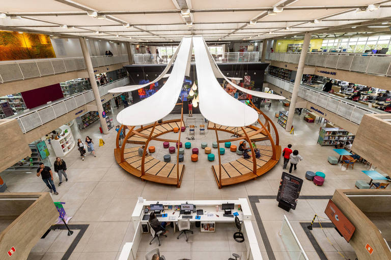 A imagem mostra o interior de uma biblioteca moderna com um design aberto. No centro, há uma estrutura em forma de círculo com assentos coloridos e uma cobertura branca. Ao fundo, prateleiras de livros e mesas de estudo são visíveis. Algumas pessoas estão caminhando e outras estão sentadas na área central. O ambiente é bem iluminado e possui um estilo contemporâneo.