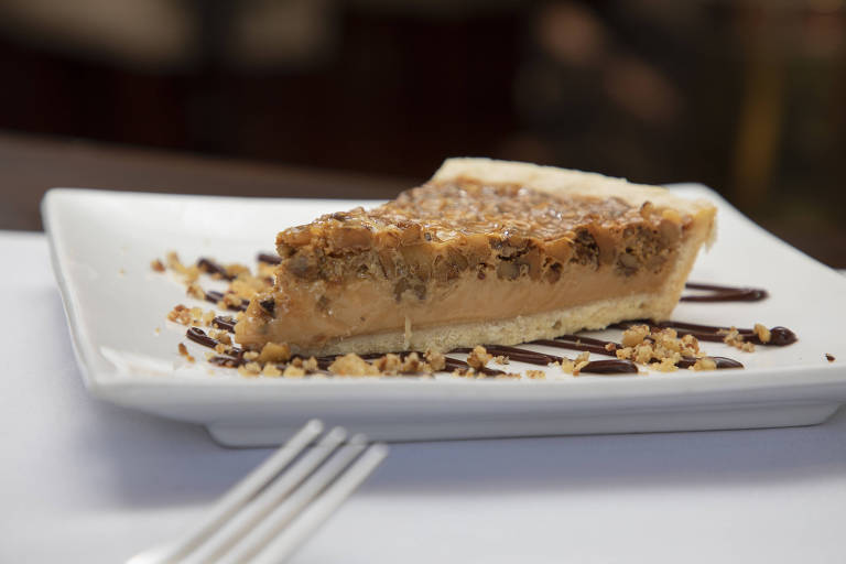 Uma fatia de torta de caramelo está servida em um prato branco. A torta tem uma crosta dourada e é coberta com uma camada de nozes picadas. Ao redor da torta， há migalhas de biscoito e um fio de calda de chocolate escuro.