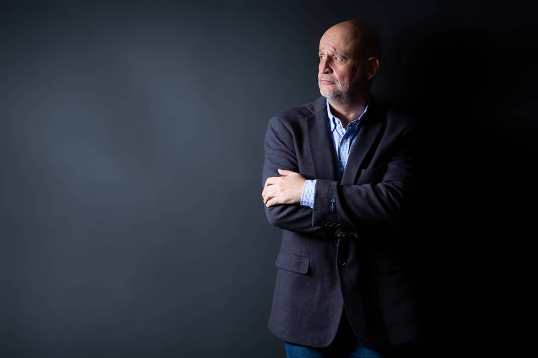 Um homem está posando em pé contra um fundo escuro. Ele está usando um blazer escuro sobre uma camisa clara e tem os braços cruzados. O homem está olhando para o lado, com uma expressão pensativa.