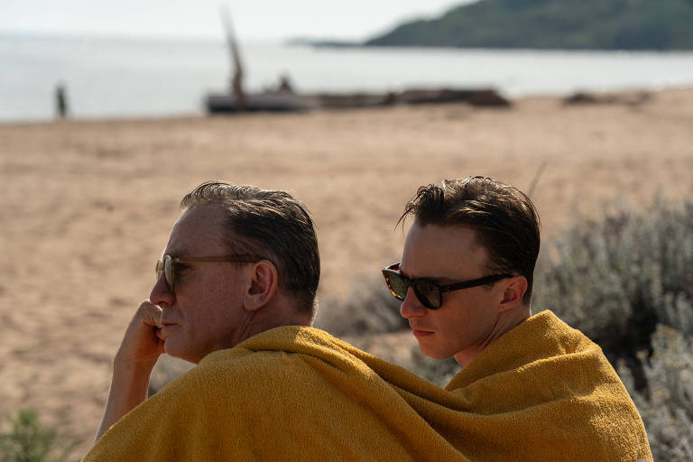 A imagem mostra dois homens sentados na praia, ambos usando toalhas amarelas. O homem à esquerda está pensativo, com a mão no queixo, e usa óculos escuros. O homem à direita está olhando para frente, também com óculos escuros. Ao fundo, há uma praia com água e vegetação, além de algumas embarcações na areia.