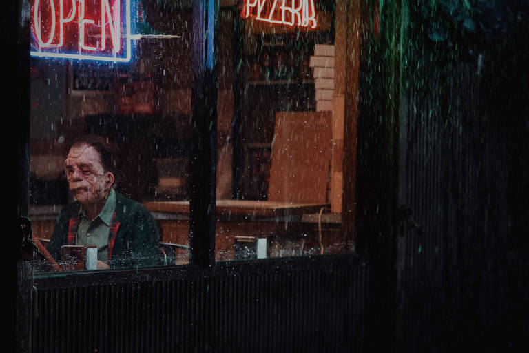 A imagem mostra um homem sentado em uma mesa dentro de uma pizzaria, visível através de uma janela embaçada pela chuva. Ele tem uma expressão pensativa e está vestido com uma camisa clara e um suéter verde. Ao fundo, há uma placa de neon 'OPEN' e outra que diz 'PIZZERIA'. A chuva cai intensamente do lado de fora, criando um efeito de gotas na janela.