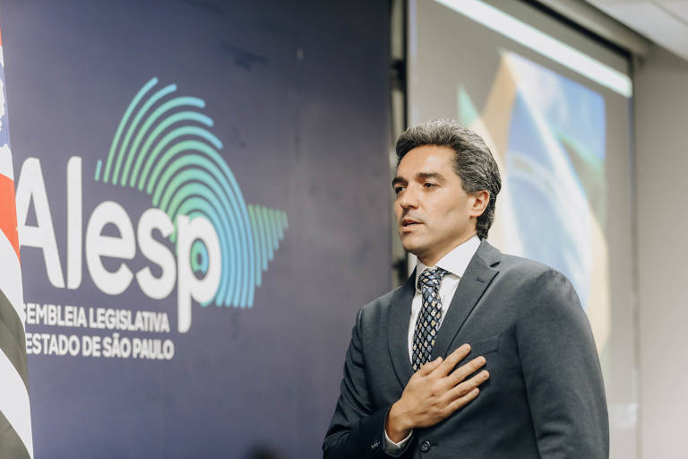 Foto mostra Paulo Mansur, um homem de pele clara em um terno preto, com camisa branca e gravata estampada com predomínio do azul-escuro. Ele está em pé, com a mão sobre o coração, em frente a um fundo que exibe o logotipo da Alesp (Assembleia Legislativa do Estado de São Paulo). Ao lado dele, há uma bandeira do estado de São Paulo.