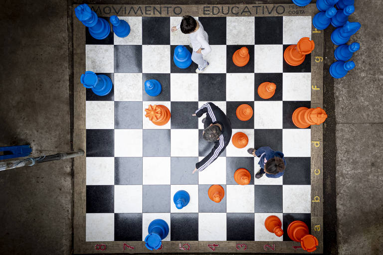 Imagem mostra um tabuleiro de xadrez gigante com peças em cores azul e laranja. Duas pessoas estão jogando， uma vestindo uma camisa branca e a outra uma camisa escura. O tabuleiro é composto por quadrados pretos e brancos， e as peças estão dispostas de maneira estratégica. Ao redor do tabuleiro， há várias peças azuis e laranjas empilhadas.