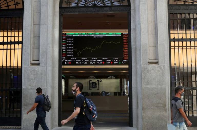 A imagem mostra a entrada de um edifício com uma tela grande exibindo gráficos e dados financeiros. Algumas pessoas estão caminhando em frente à entrada, que possui portas de vidro e uma grade. A tela apresenta informações sobre o mercado financeiro, incluindo gráficos de alta.