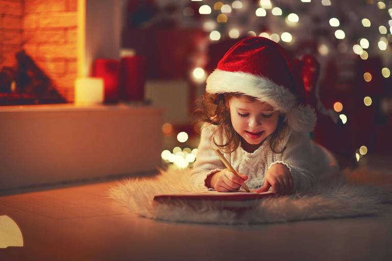 Menina com gorro de Papai Noel deitada no chão escrevendo carta com árvore de Natal iluminada ao fundo