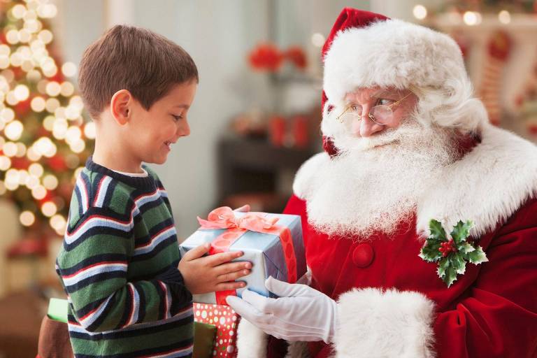 Menino recebendo presente de homem vestido de Papai Noel com decoração de Natal ao fundo