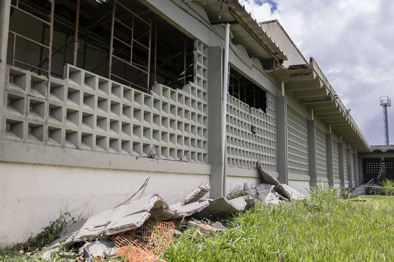 A imagem mostra um edifício com paredes de blocos de concreto, algumas janelas estão sem vidro e há uma estrutura de andaimes visível. Na parte inferior, há destroços de concreto e uma área de grama ao redor. O céu está parcialmente nublado.