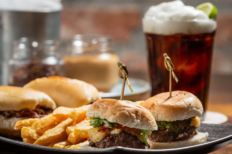 A imagem mostra um prato com dois pequenos hambúrgueres, cada um com um palito, acompanhados de batatas fritas. Ao fundo, há um copo de bebida escura com espuma e uma fatia de limão. Também são visíveis alguns potes com ingredientes em conserva.