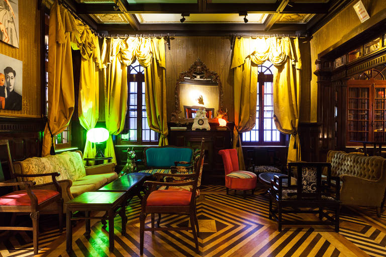 A imagem mostra um bar decorado com um estilo vintage. As paredes são de um tom escuro, e há janelas com cortinas amarelas. O piso é de madeira com um padrão de listras. Há vários móveis, incluindo sofás e cadeiras coloridas, além de mesas de centro. A iluminação é suave, com lâmpadas de diferentes cores.