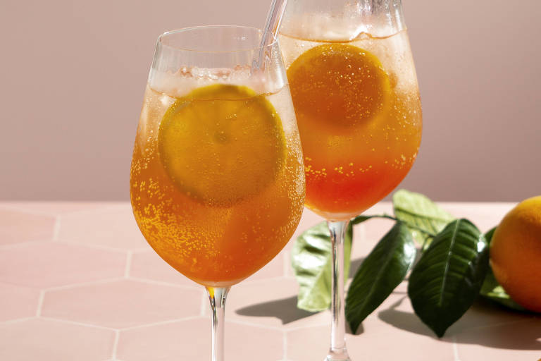A imagem mostra dois copos de bebida com gás， decorados com fatias de laranja， em uma mesa de superfície rosa. Os copos têm canudos transparentes e estão acompanhados de folhas verdes.