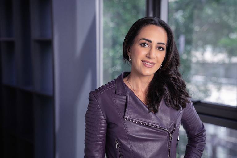 A imagem mostra uma mulher sorrindo, vestindo uma jaqueta de couro roxa com detalhes em relevo. Ela está posicionada em um ambiente interno, próximo a uma janela que permite a entrada de luz natural. Ao fundo, há prateleiras escuras. A mulher tem cabelo longo e solto, e usa brincos. O ambiente é iluminado e transmite uma sensação de modernidade.
