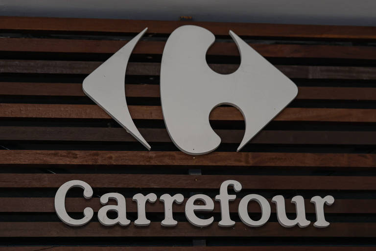 A imagem mostra o logotipo da rede de supermercados Carrefour. O logotipo é composto por um símbolo que se assemelha a um 039;C039; estilizado， com duas setas apontando para fora， e abaixo está escrito 039;Carrefour039; em letras minúsculas， em uma fonte clara e simples. O fundo é de madeira com ripas horizontais.