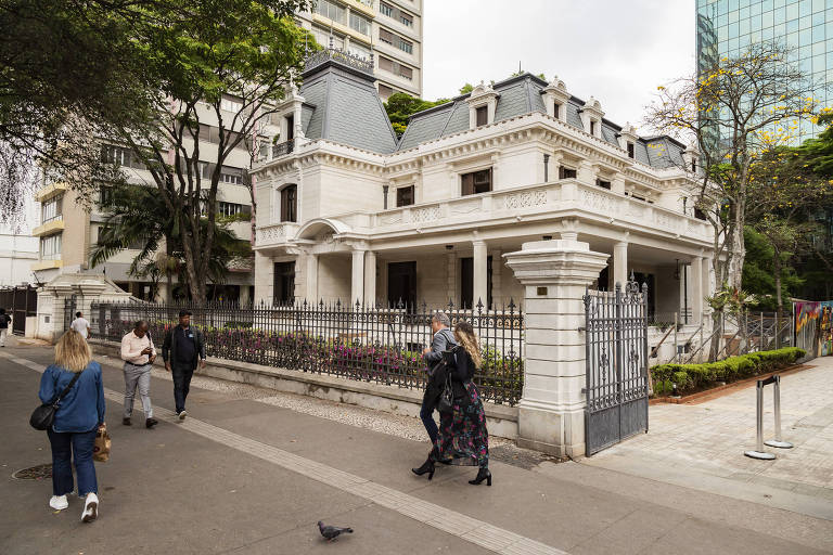 A imagem mostra uma mansão de estilo arquitetônico clássico, com paredes brancas e telhados de duas águas. A construção possui detalhes ornamentais e janelas com grades. Em frente à mansão, há um passeio onde várias pessoas caminham, incluindo uma mulher com cabelo loiro e uma saia longa floral. Um homem de camisa clara e outro de calça escura estão próximos. Um pombo está pousado no chão. Ao fundo, há prédios modernos e árvores.