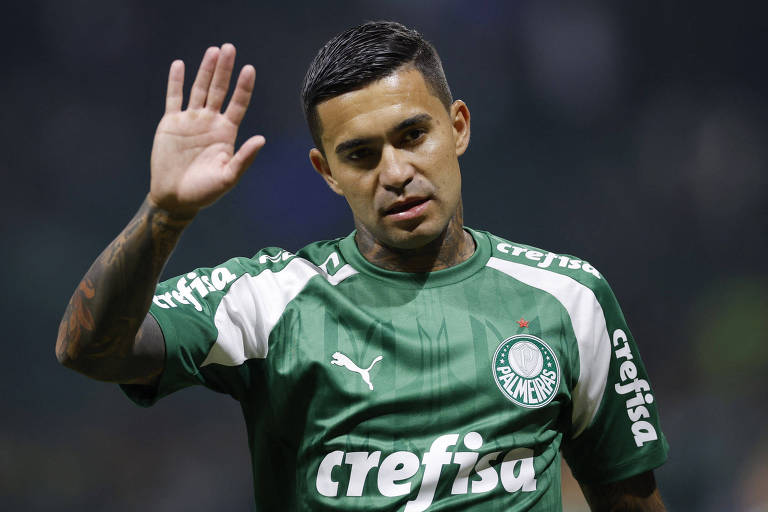 Um jogador de futebol está acenando com a mão direita. Ele veste um uniforme verde com detalhes brancos e o logotipo de um patrocinador. O fundo é desfocado， sugerindo um ambiente de estádio.
