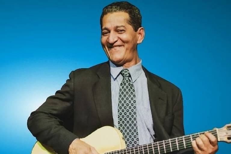 Um homem sorridente está sentado segurando um violão. Ele usa um terno escuro, uma camisa clara e uma gravata com padrão. O fundo da imagem é azul, criando um contraste com sua roupa. O homem parece estar se divertindo enquanto toca o instrumento.