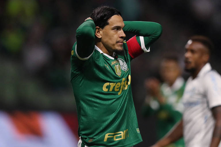 Foto mostra o zagueiro paraguaio Gustavo Gómez em campo durante uma partida do campeonato brasileiro contra o Grêmio. A imagem registra o atleta de pele clara da cintura para cima， ele veste a camisa verde do Palmeiras， com a segunda pele verde por baixo da camiseta. Ele esta com as duas mãos na cabeça e e olha fixamente para o lado direito da imagem. Ele tem longos cabelos pretos e lisos que estão seguros por uma bandana preta. Ele tem um cavanhaque preto. Na manga do braço esquerdo do zagueiro está uma grossa faixa vermelha. Que indica que ele é capitão do time. O fundo da imagem é escuro， mas pela posição da foto são torcedores nas arquibancadas.  