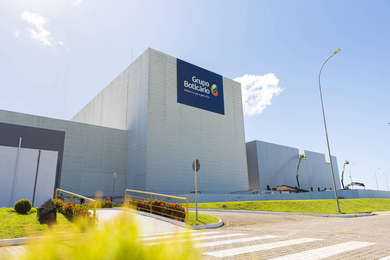 Imagem de uma grande estrutura industrial com uma fachada moderna. O edifício é de cor clara e possui um grande letreiro com o nome da empresa. O céu está limpo e azul, e há algumas nuvens brancas. Na frente do edifício, há uma área verde com grama e arbustos. Também é visível um poste de luz e uma estrada pavimentada.