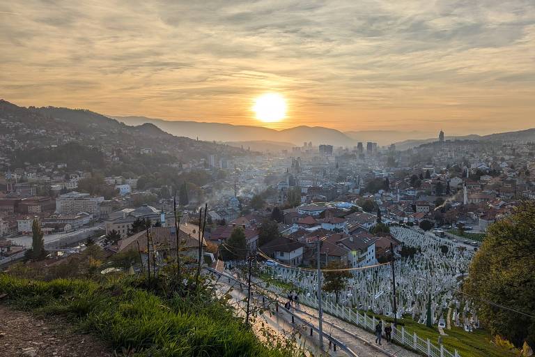 A imagem mostra uma vista panorâmica da cidade de Sarajevo ao pôr do sol. No primeiro plano， há um cemitério com lápides brancas， enquanto a cidade se estende abaixo， com casas de telhados vermelhos e edifícios modernos. O sol está se pondo atrás das montanhas， criando um céu com nuvens e tons alaranjados. A vegetação é visível nas bordas da imagem， e algumas pessoas estão caminhando na estrada que desce em direção à cidade.