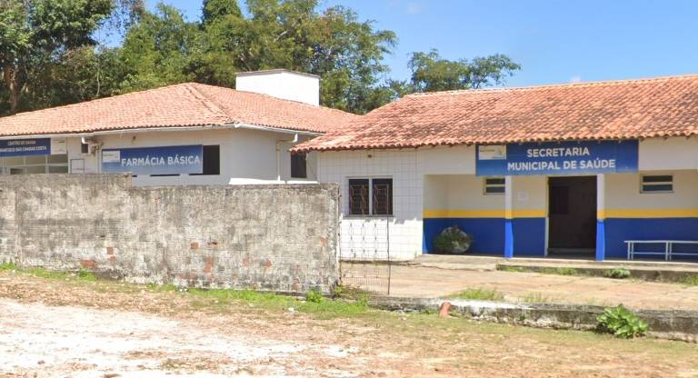 A imagem mostra um prédio com duas seções: à esquerda, a 'FARMÁCIA BÁSICA' e, à direita, a 'SECRETARIA MUNICIPAL DE SAÚDE'. O edifício tem um telhado de duas águas e é cercado por um muro. O ambiente é claro, com vegetação ao fundo e um espaço de terra na frente.