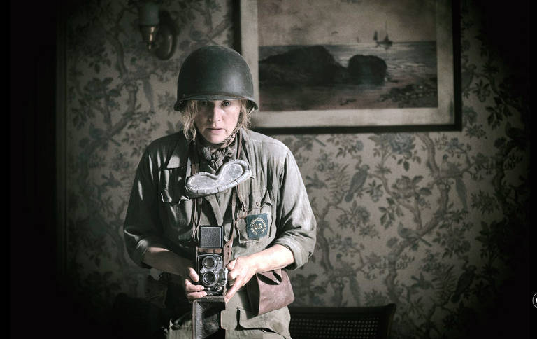 A imagem mostra uma mulher vestindo um uniforme militar e um capacete, segurando uma câmera fotográfica. Ela está em um ambiente interno, com uma parede decorada com papel de parede floral e uma pintura de paisagem ao fundo. A mulher tem uma expressão séria e parece estar focada. O uniforme possui um emblema que diz 'U.S.' e um acessório em forma de coração ao redor do pescoço.