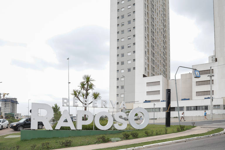 A imagem mostra um letreiro grande com a palavra 'RESERVA RAPOSO' em letras brancas, localizado em uma área urbana. Ao fundo, há um edifício alto e moderno, com janelas e uma fachada predominantemente branca. O ambiente é urbano, com algumas árvores e um gramado na frente do letreiro. O céu está nublado.