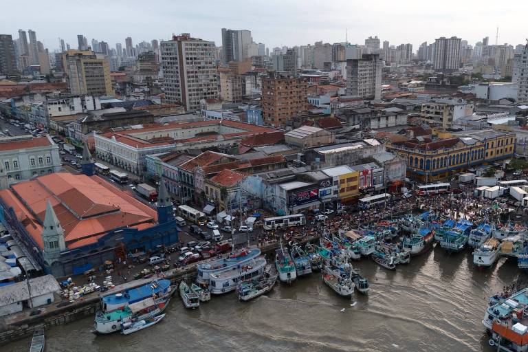 A imagem mostra uma vista aérea de um porto urbano， com vários barcos ancorados em um canal. Ao fundo， há uma densa área urbana com prédios altos e uma grande quantidade de estruturas. O céu está nublado， e a água do canal reflete a luz. Há também uma movimentação de pessoas e veículos nas proximidades do porto.