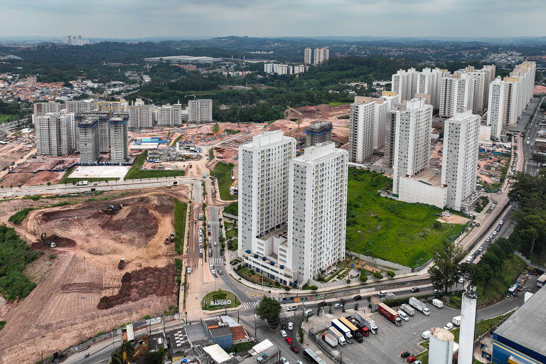 A imagem mostra uma vista aérea de uma área urbana em construção, com vários edifícios altos e novos em primeiro plano. No fundo, há terrenos vazios e em obras, além de áreas verdes. O céu está nublado, e a paisagem urbana é marcada por ruas e veículos visíveis na parte inferior da imagem.