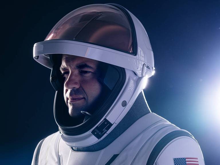 A imagem mostra um astronauta usando um traje espacial branco com detalhes em cinza. O capacete é transparente e reflete a luz. O astronauta está de perfil， com uma expressão séria， e há uma bandeira dos Estados Unidos no ombro esquerdo do traje. O fundo é escuro， com luzes azuis e brancas que criam um efeito dramático.