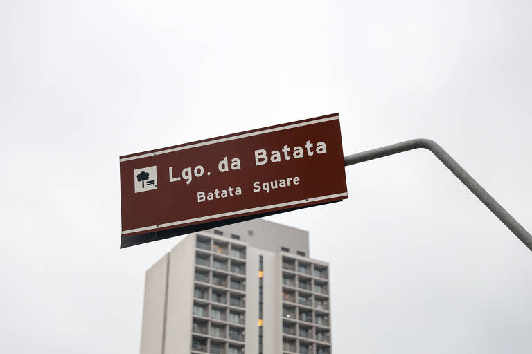 A imagem mostra uma placa de sinalização marrom com o texto 'Lgo. da Batata' e 'Batatas Square'. Ao fundo, há um edifício residencial de vários andares. O céu está nublado.