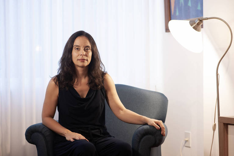 Uma mulher com cabelo longo e solto está sentada em um sofá azul. Ela usa um vestido preto e está em um ambiente iluminado, com uma lâmpada ao lado e uma cortina branca ao fundo.