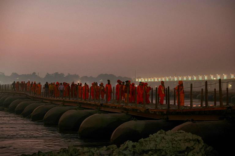 A imagem mostra um grupo de pessoas vestidas com roupas laranjas caminhando sobre uma estrutura flutuante, provavelmente um caminho de madeira, sobre a água. O cenário é ao amanhecer, com um céu em tons de rosa e azul. Ao fundo, há uma ponte iluminada e árvores, enquanto a água reflete a luz suave do ambiente. Algumas pessoas estão paradas, enquanto outras estão em movimento.