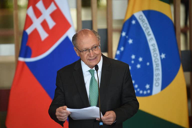 Um homem de terno escuro e gravata verde está falando ao microfone, segurando um papel. Ao fundo, há bandeiras da Eslováquia e do Brasil. O ambiente parece ser um evento oficial, com uma iluminação suave.