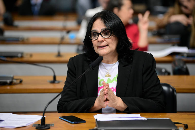 Damares Alves， uma mulher com cabelo longo e preto， usando óculos e um blazer preto， está sentada à mesa em uma audiência pública. Ela parece estar falando ou respondendo a perguntas. Ao fundo， há outras pessoas， algumas levantando as mãos. A mesa tem papéis e um celular visíveis.