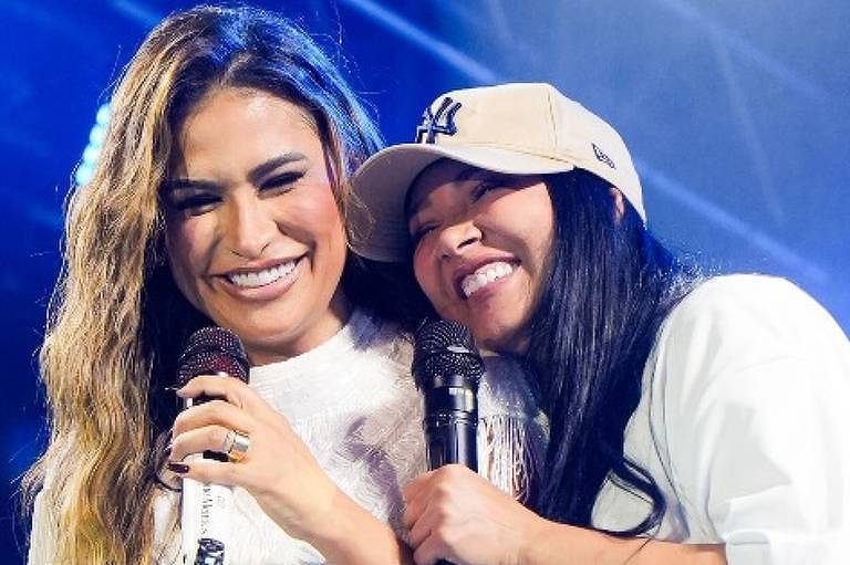 Em foto colorida, duas mulheres cantam juntas em cima de um palco