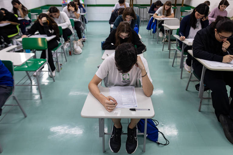 Foto mostra jovens sentados em cadeiras com cabeças baixas lendo provas em uma sala de aula