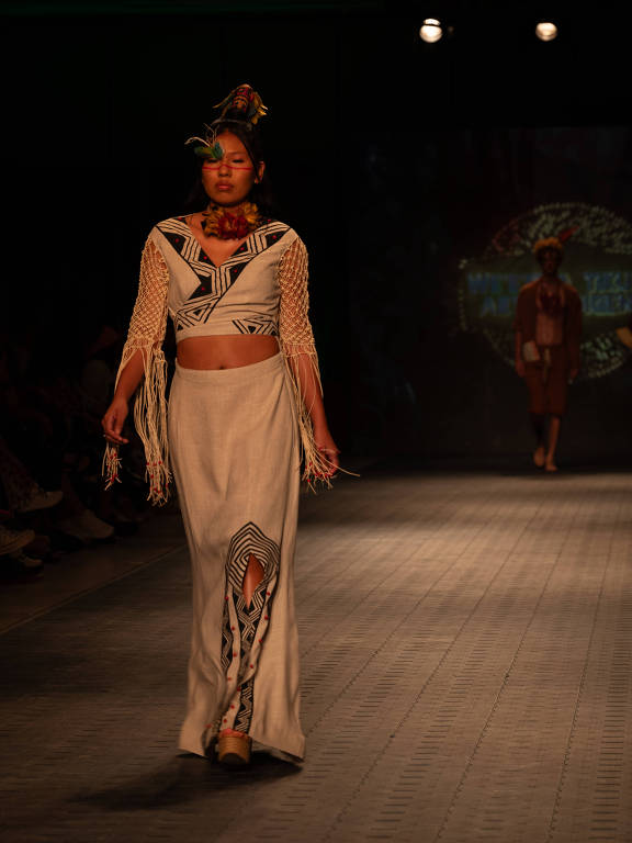 Uma modelo caminha na passarela usando um conjunto de roupas com temática indígena. A blusa é de manga longa com detalhes em crochê e um padrão geométrico em preto e branco. A saia longa possui um recorte e também apresenta padrões semelhantes. A modelo tem adornos de penas na cabeça e colares coloridos, além de pintura facial em tons de vermelho e verde. Ao fundo, há uma tela com iluminação que complementa o ambiente do desfile.