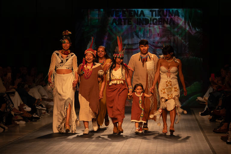 Um grupo de pessoas caminha em uma passarela durante um desfile de moda indígena. Eles estão vestidos com trajes tradicionais, incluindo saias e vestidos feitos de materiais naturais. Algumas pessoas usam adornos na cabeça, como penas e colares coloridos. Ao fundo, há uma tela com a inscrição 'WELENA TIKUNA ARTE INDÍGENA'. O público aplaude e observa atentamente.