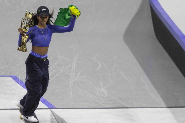 Uma skatista está em uma prancha de skate, segurando um troféu dourado em uma mão e uma bandeira verde e amarela na outra. Ela usa uma blusa de manga longa azul e calças largas. O fundo é uma pista de skate com superfícies em tons de cinza e azul.